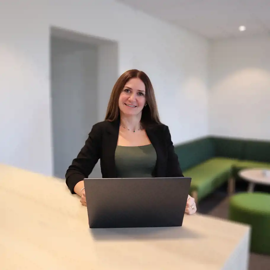 Leende kvinna i svart kavaj som sitter vid en bärbar dator i en modern kontorsmiljö med gröna soffor i bakgrunden. Joumana Maghamez är rådgivare på Almi.