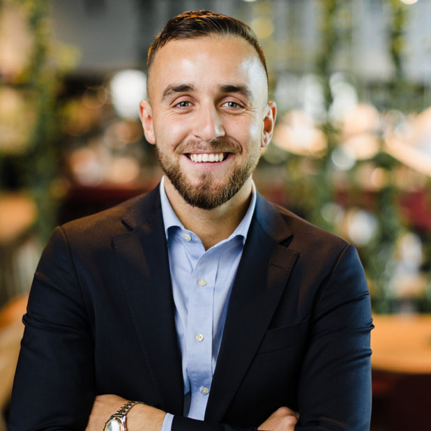 Leende man i kostym som sitter med armarna i kors i en modern och livlig kontorsmiljö med gröna växter i bakgrunden. Adam Silverlid är rådgivare på Almi.