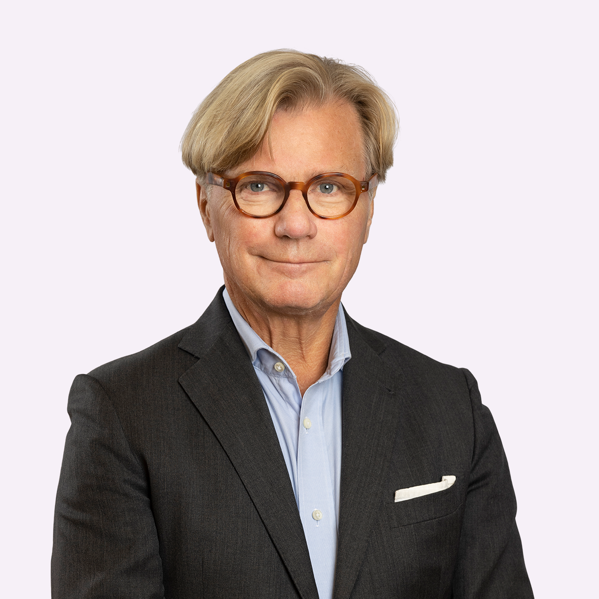 Porträttbild av en man i kostym och ljusblå skjorta. Han har runda glasögon med en brun båge och en vänlig, självsäker uppsyn. Bakgrunden är ljus och diskret, vilket ger en stilren och professionell framtoning. Bilden passar utmärkt för affärssammanhang eller officiella presentationer.