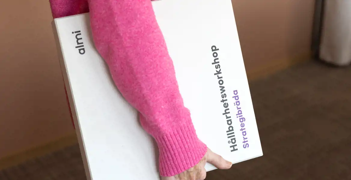 En person i rosa tröja håller i en vit låda med texten "Hållbarhetsworkshop Strategibräda" och logotypen för Almi. Bakgrunden är inomhusmiljö med en beige vägg.