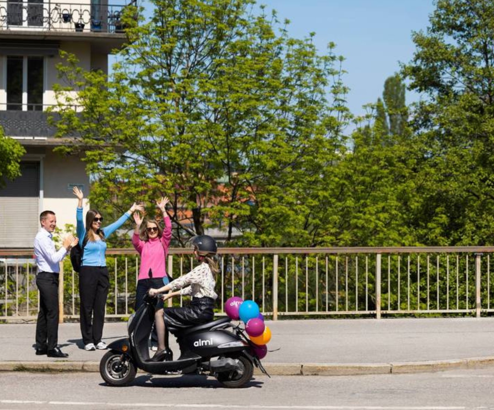 kvinna kör moped med ballonger baktill och kör förbi 3 personer vid vägen som vinkar glatt