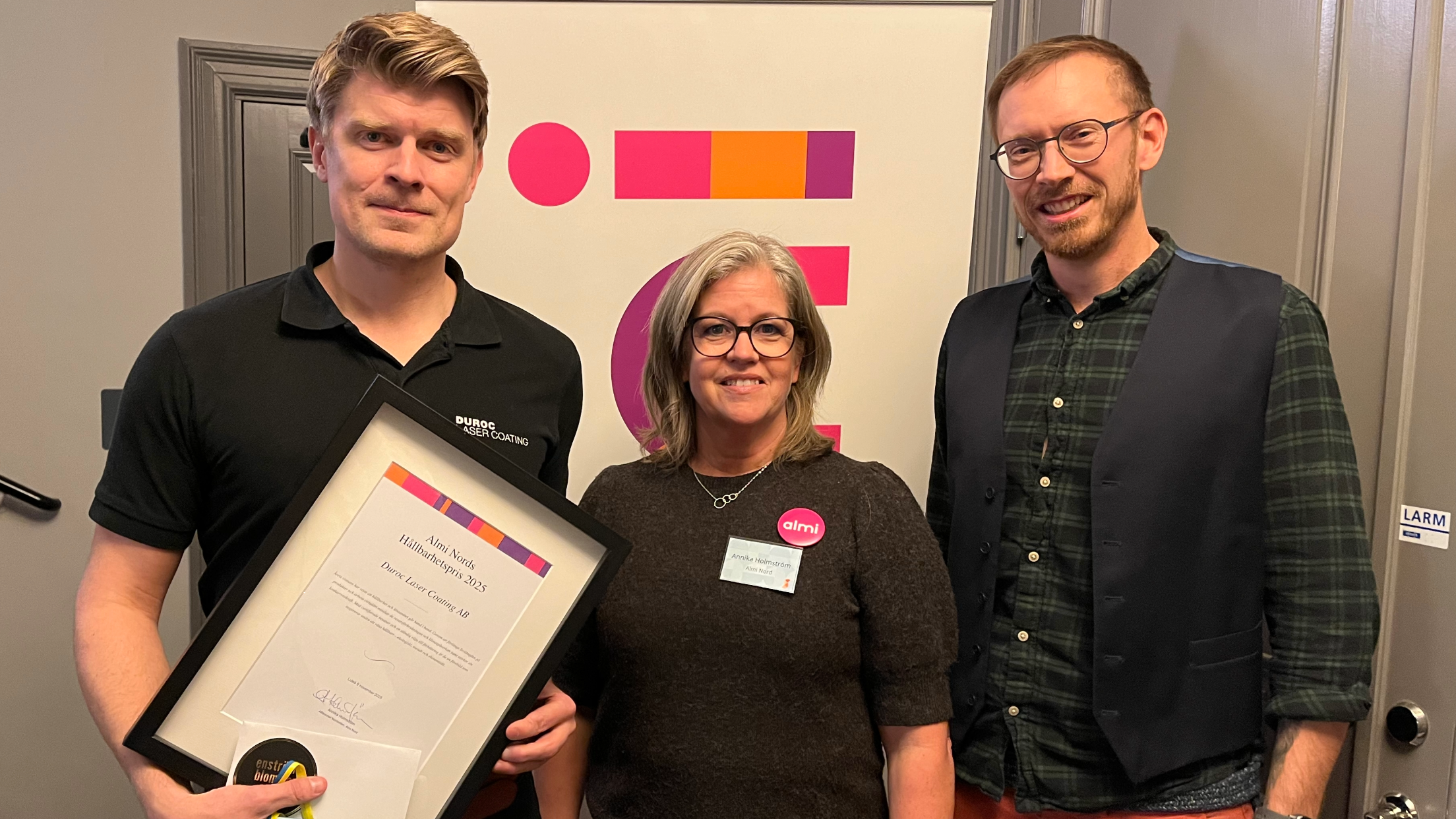 Tre personer står framför en affisch med färgglada geometriska former. Den vänstra personen håller ett inramat diplom för Hållbarhetspris från Almi.