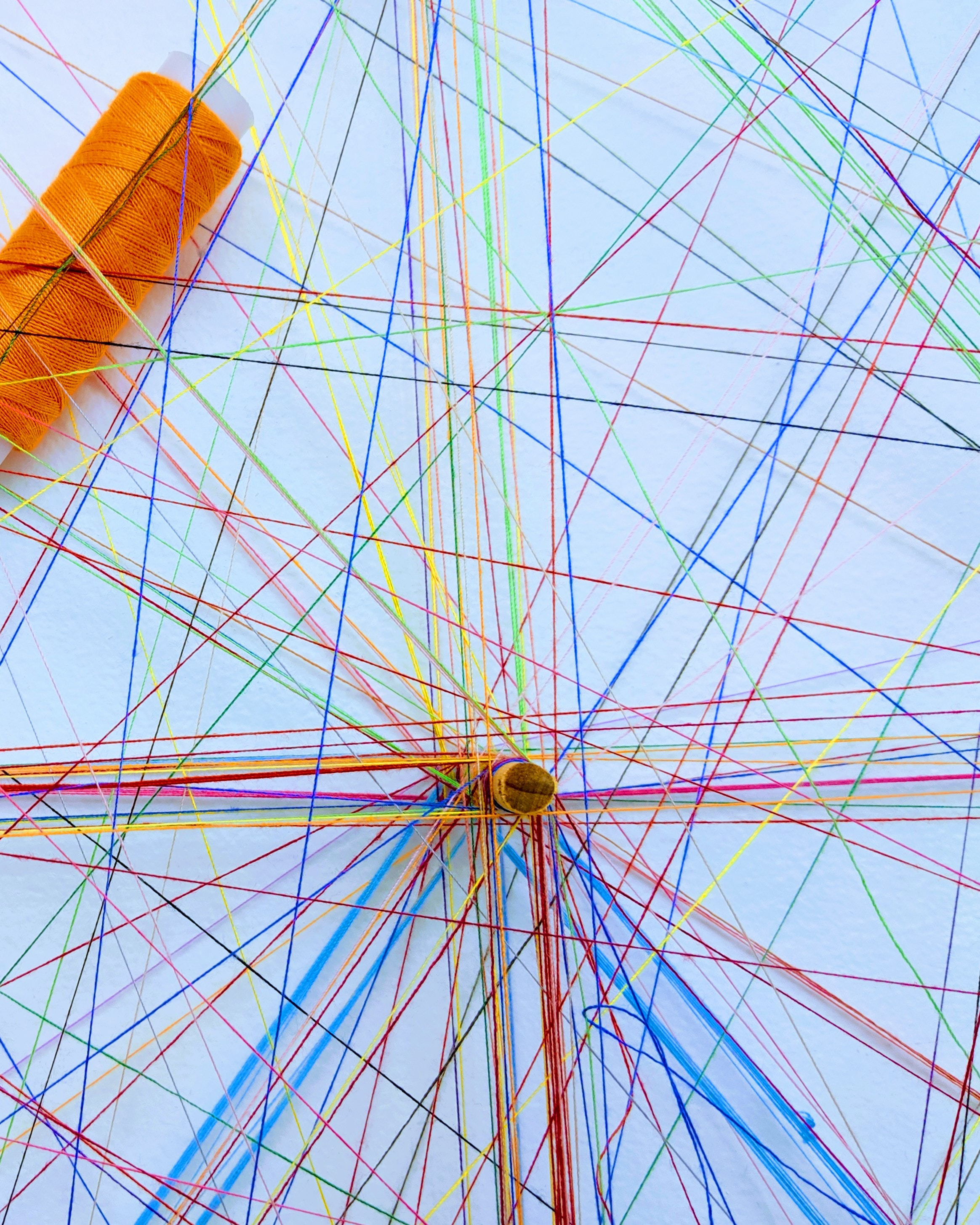 En konstnärlig installation med färgglada trådar spända mellan olika punkter på en vit bakgrund, med en orange trådrulle placerad i hörnet.