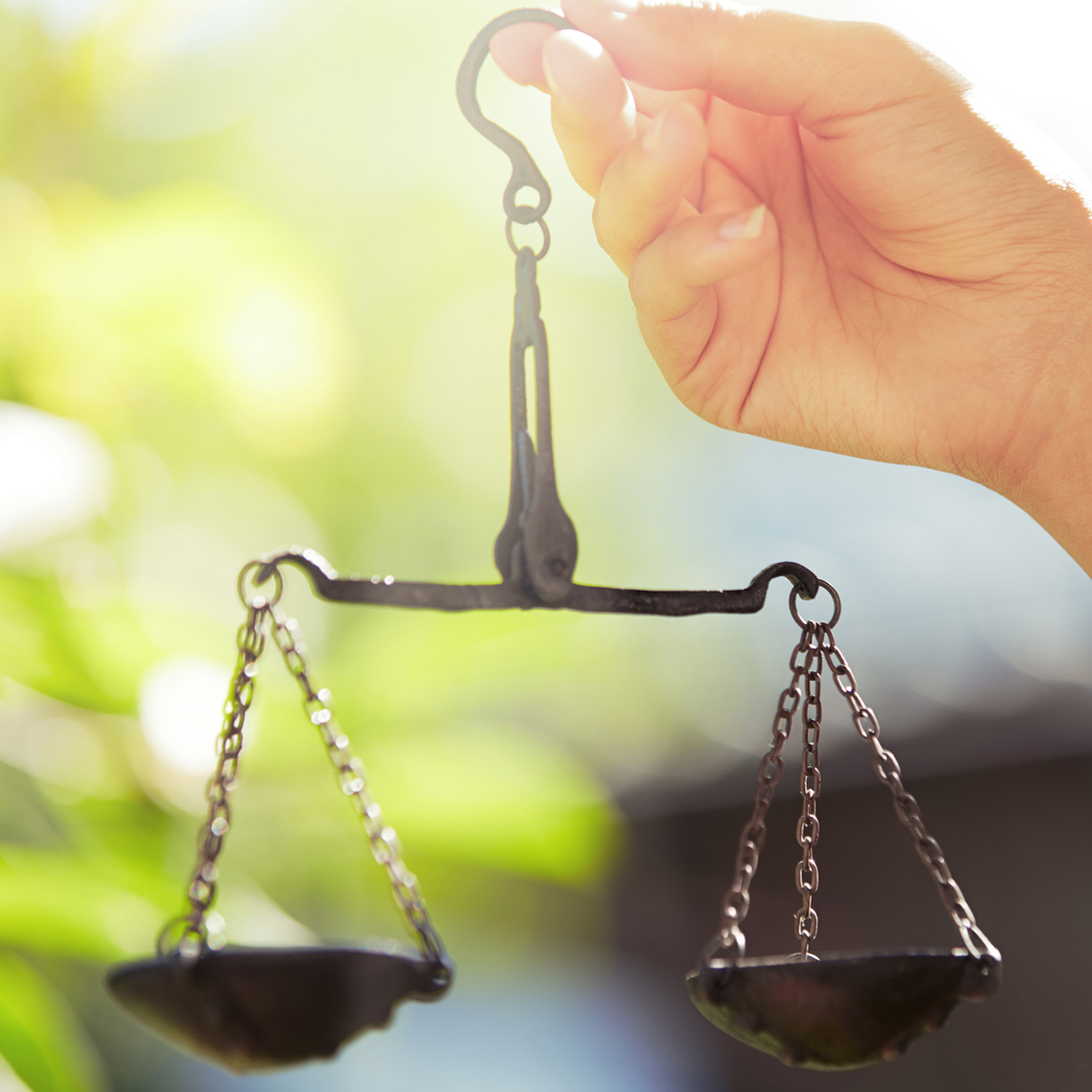En hand håller upp en liten, klassisk balansvåg i smide, med två kedjeförsedda vågskålar. Bakgrunden är suddig med gröna blad och solljus.
