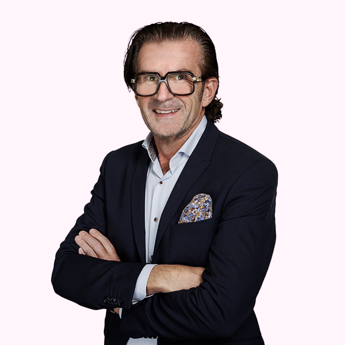 Patrik Kinnbom, Investment Manager på Almi Invest Småland och öarna, står leende med armarna i kors, klädd i mörkblå kavaj, ljus skjorta och mönstrad näsduk mot en ljusrosa bakgrund.