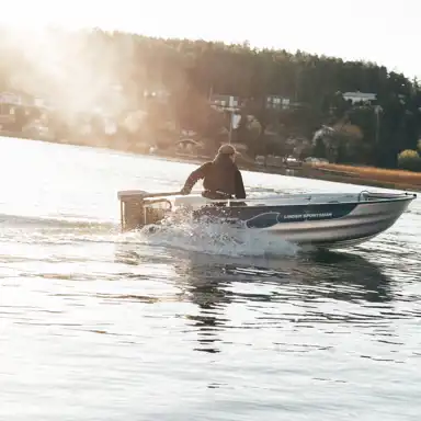 En person kör en aluminiumbåt med Zpark-elutombordare över lugnt vatten i solnedgång. På båtsidan står det "Linder Sportsman". Hus och skog syns i bakgrunden.