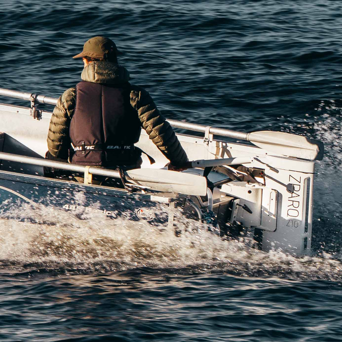 En person kör en liten motorbåt på öppet vatten. Personen bär grön keps, grön dunjacka och flytväst. Båten har en elektrisk utombordsmotor från Zpark, modell Z10. Vattnet krusar sig bakom båten i soligt väder.
