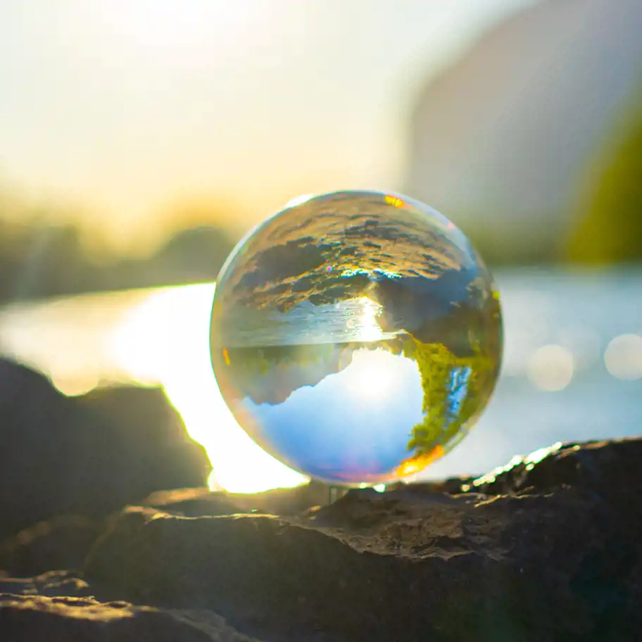 A glass sphere placed on a rocky surface by a sunlit riverbank, reflecting an upside-down image of the surrounding landscape with bright sunlight, blue sky, clouds, and greenery.